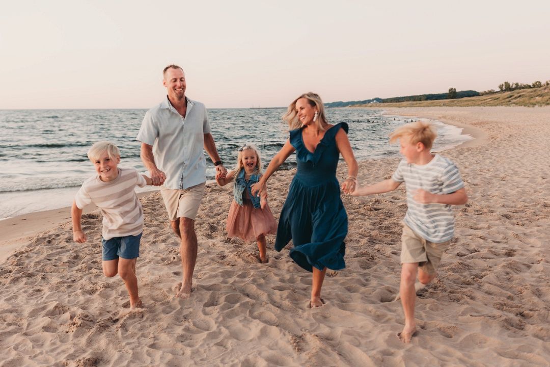 A joyful family of five runs and plays together along a sunny sandy beach at sunset.
