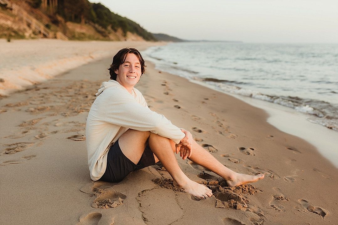 Young person sitting barefoot on sandy beach smiling, golden hour light, waves in background.