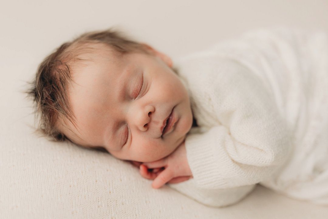 Peaceful sleeping newborn baby in white knit outfit lying on soft cream blanket, smiling slightly.