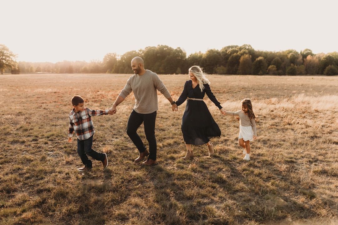Family of four holding hands walking through a golden field at sunset.