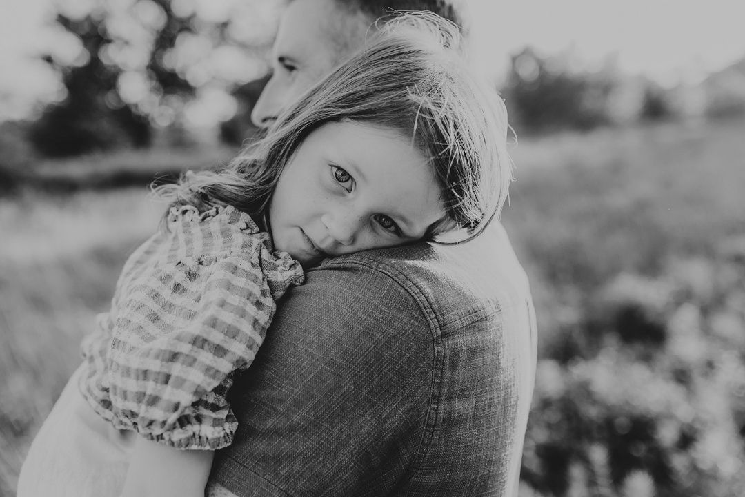A young girl rests her head on a mans shoulder in a tender black and white outdoor portrait.