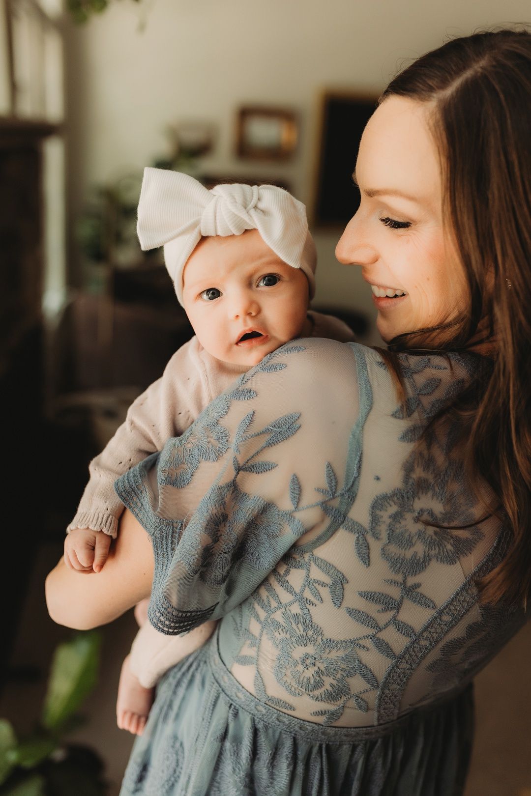 A smiling mother holds her baby girl with a white bow headband indoors.