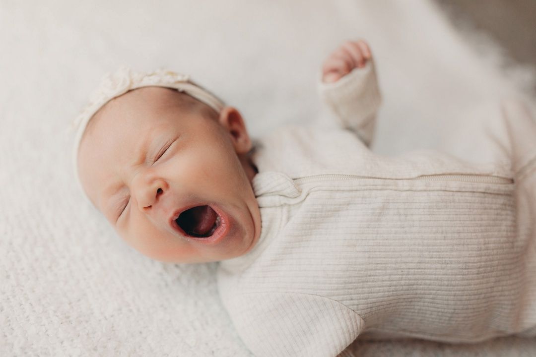Adorable newborn baby in cream knit outfit yawning widely while lying on soft white blanket.