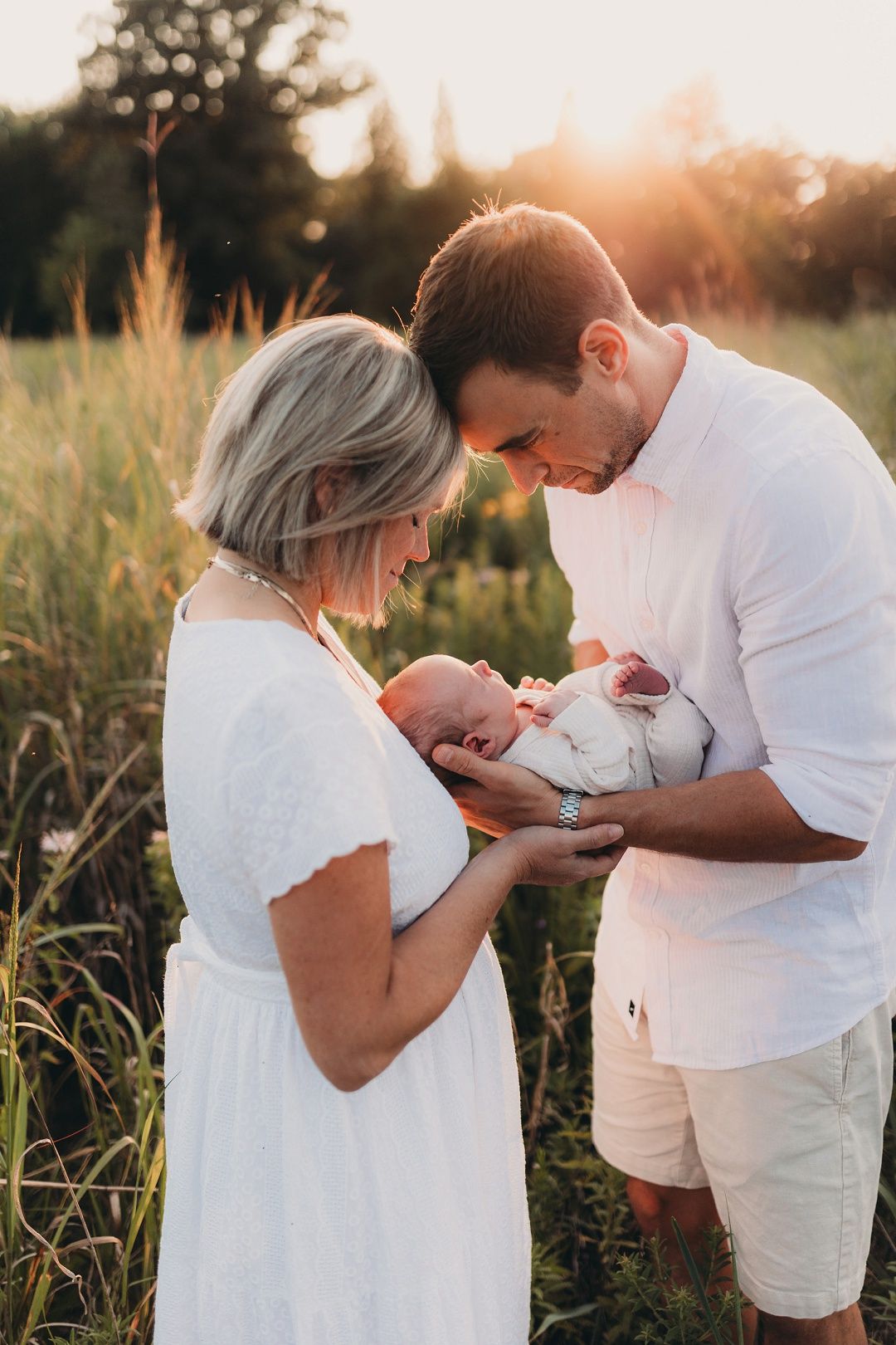Parents dressed in white admire their newborn baby during a golden sunset outdoors.