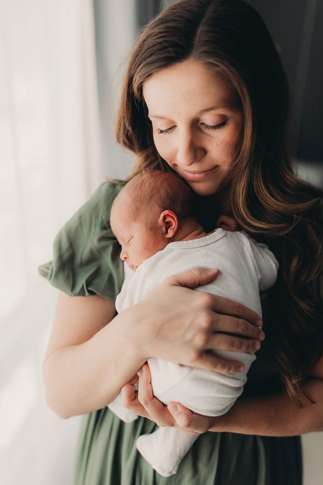 Dark-haired mother in green dress tenderly holding sleeping newborn baby near bright window.