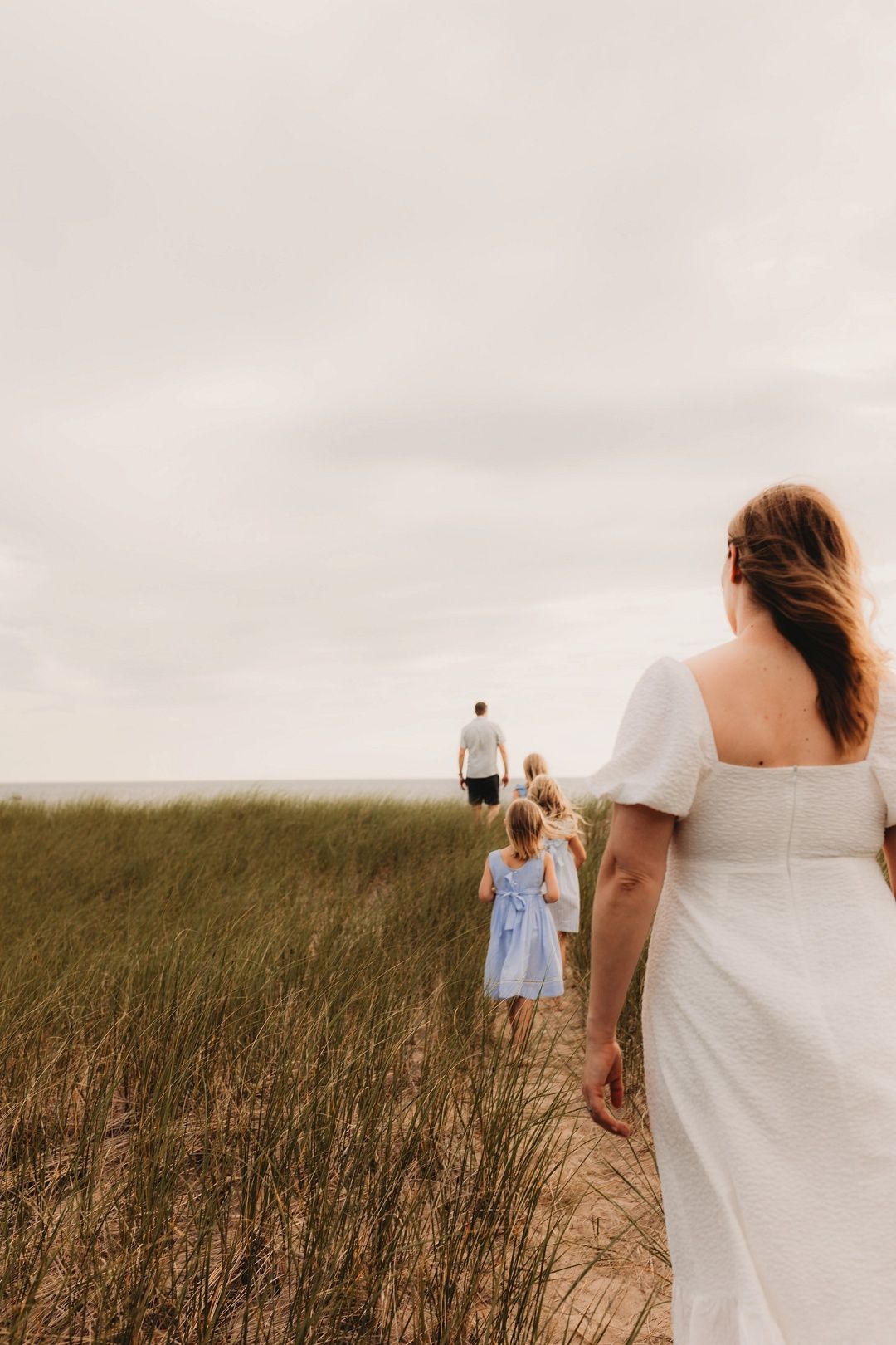 Family walking through beach grass dunes toward the ocean on an overcast day.