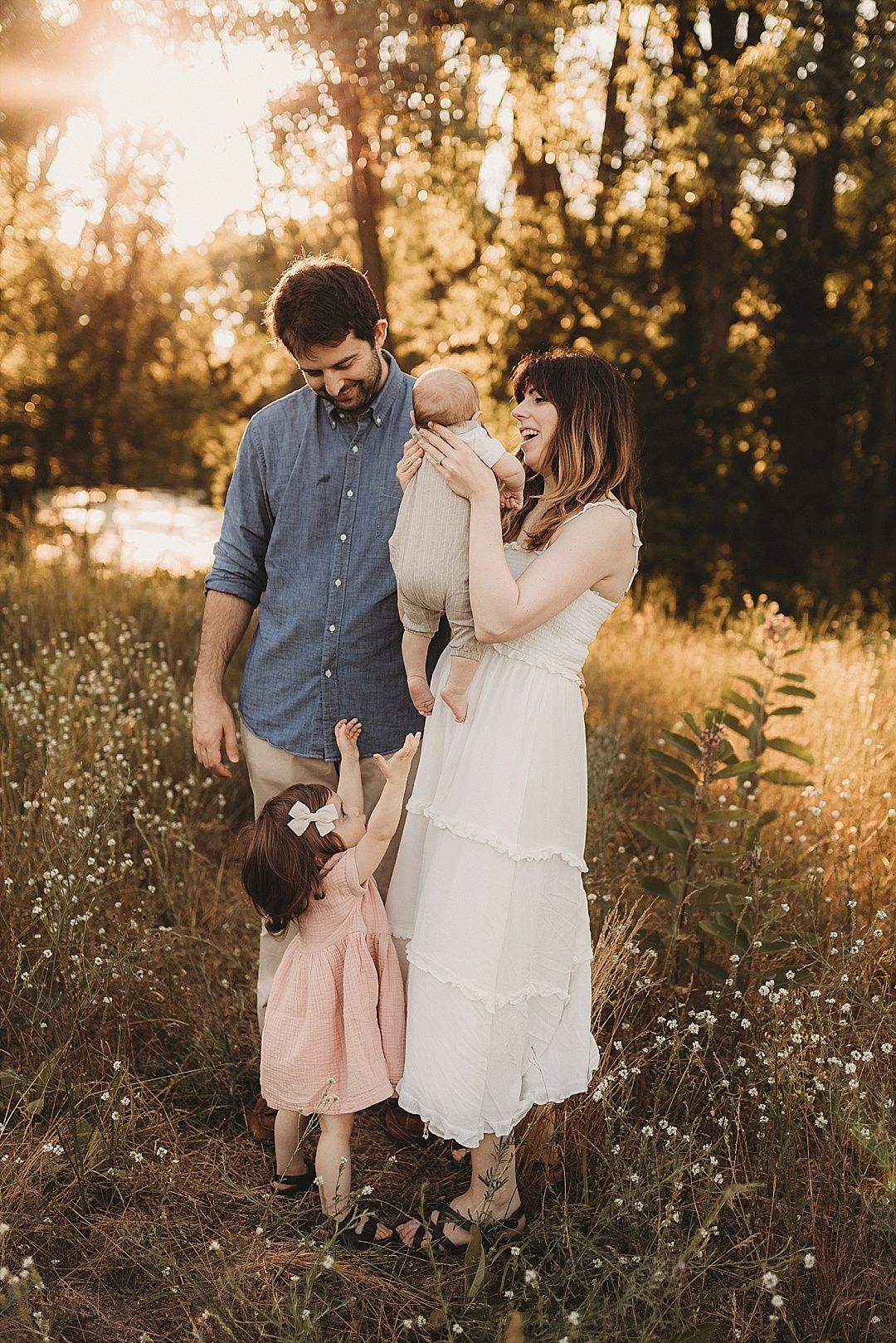 A happy family with a toddler girl and newborn baby poses in a golden sunlit meadow.