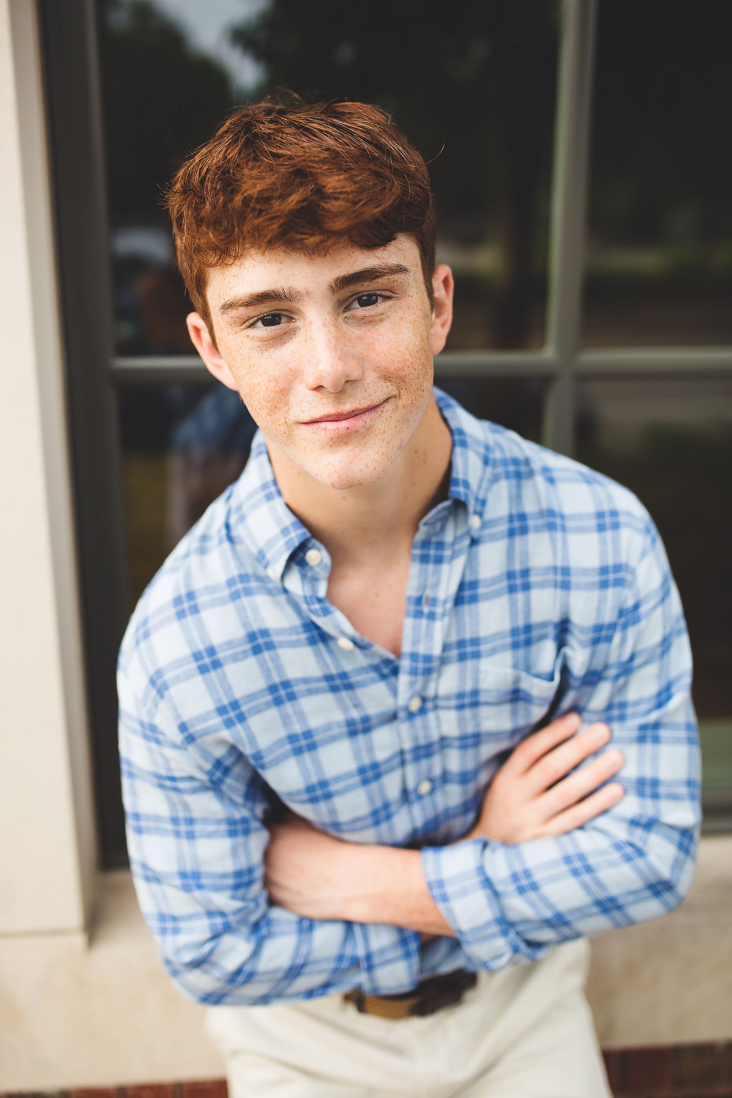 Red-haired teenage boy in blue plaid shirt standing with arms crossed, smiling confidently.