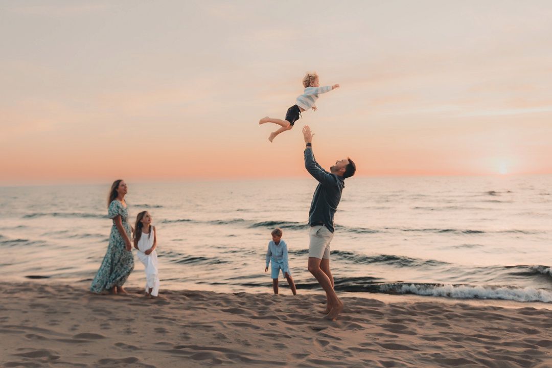 A father joyfully tosses a toddler in the air on a beach at sunset with family nearby.