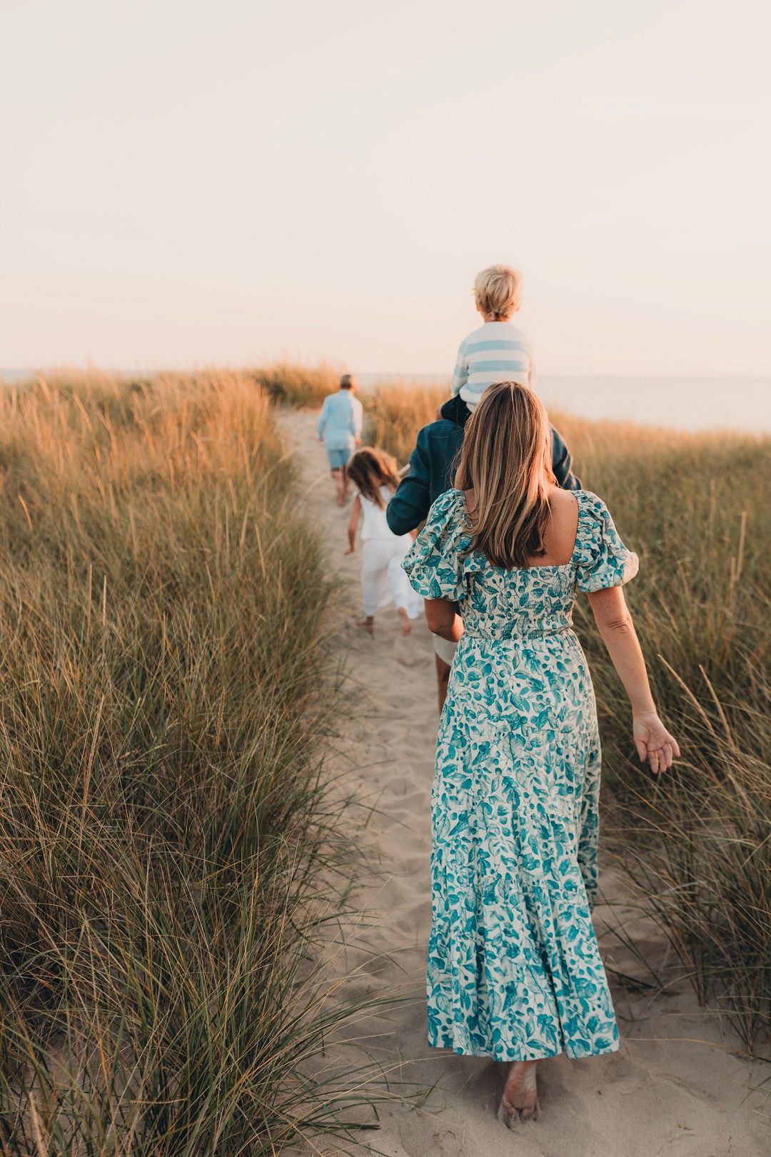 Family walking on sandy beach path through tall grass at golden sunset hour.