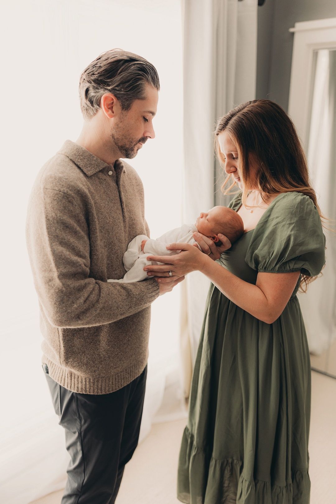 New parents in neutral tones lovingly admiring their newborn baby together near bright window.