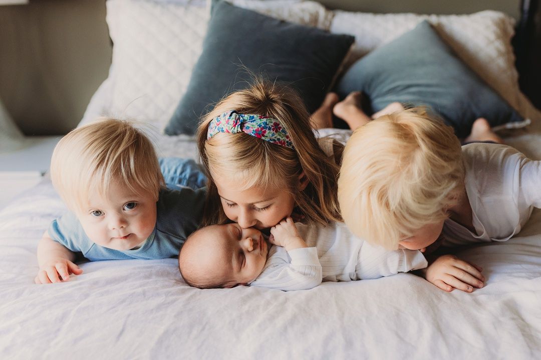 Three young siblings lie on a bed lovingly surrounding their newborn baby.