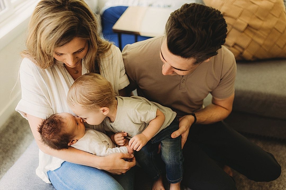 A loving family of four sits together on the floor as older sibling meets the new baby.