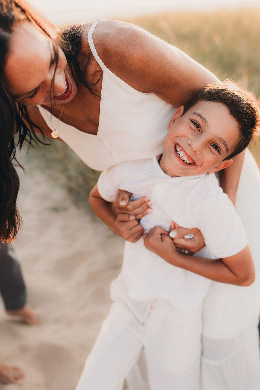 A laughing boy is hugged from behind by his smiling mother in a sunny outdoor setting.
