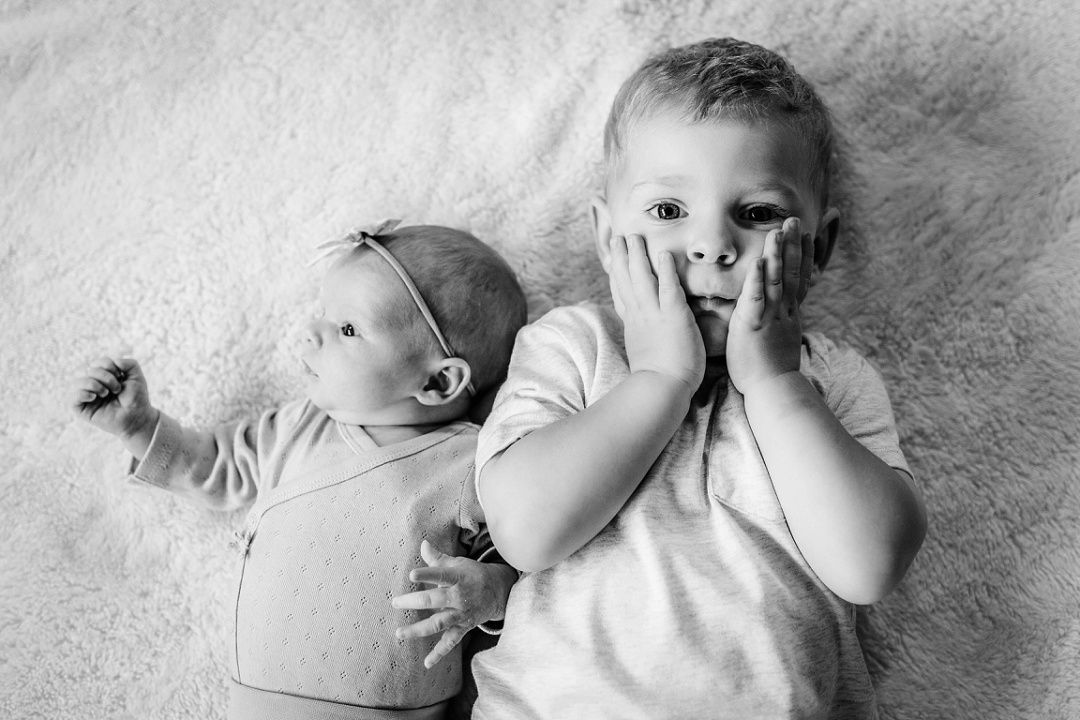 Black and white photo of toddler lying next to newborn sibling on soft blanket.