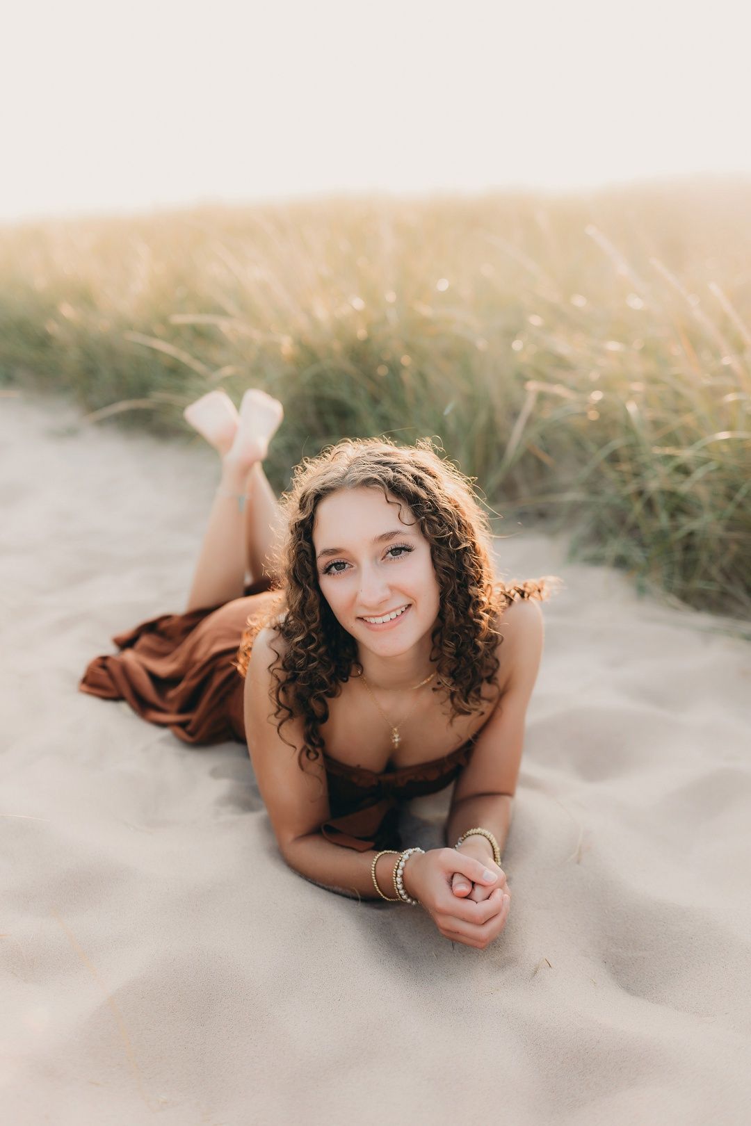 Smiling teen girl with curly hair in brown dress lying on sandy beach dunes for senior portraits.