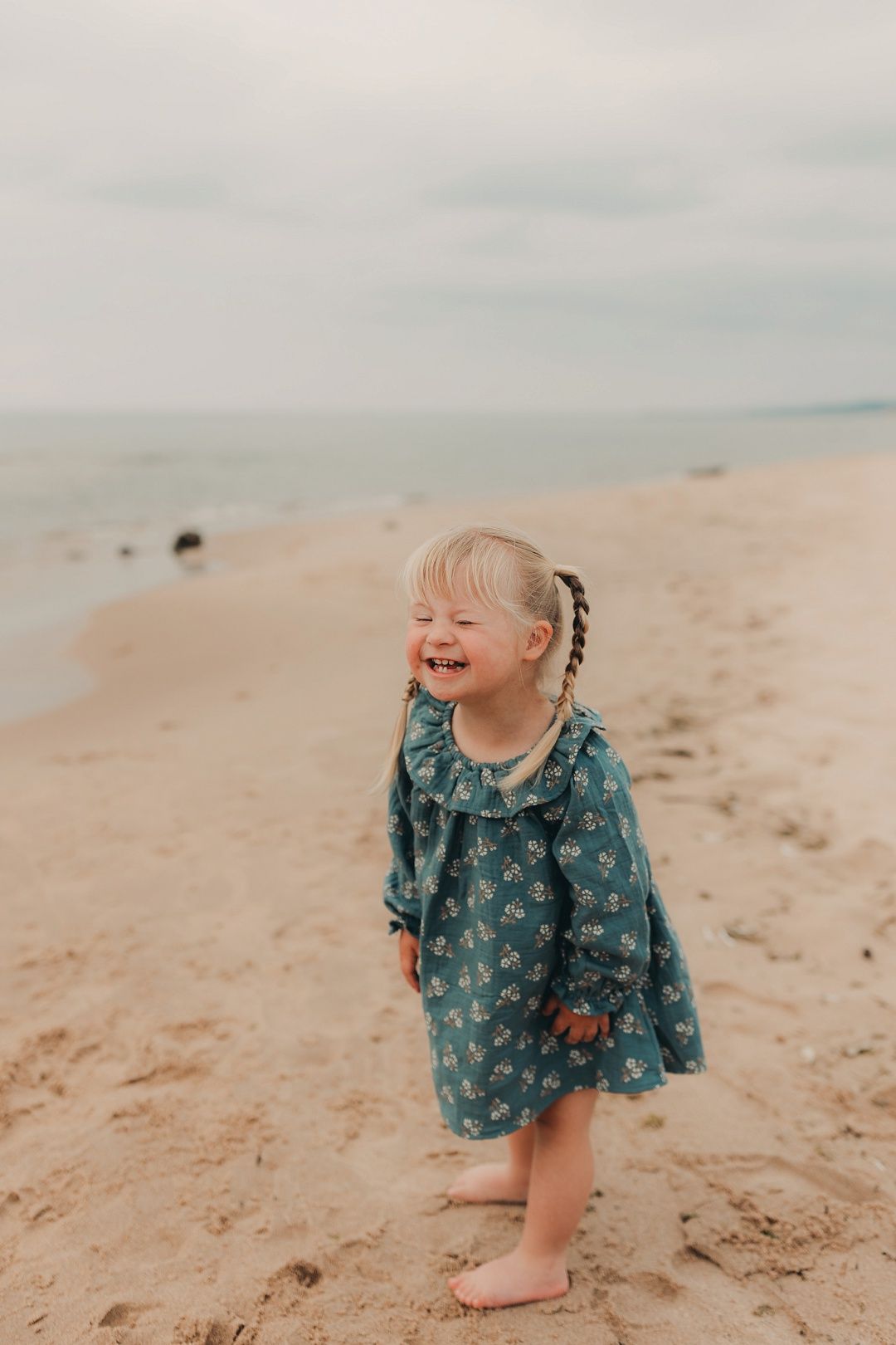 Adorable toddler girl in teal floral dress laughs joyfully on sandy beach.