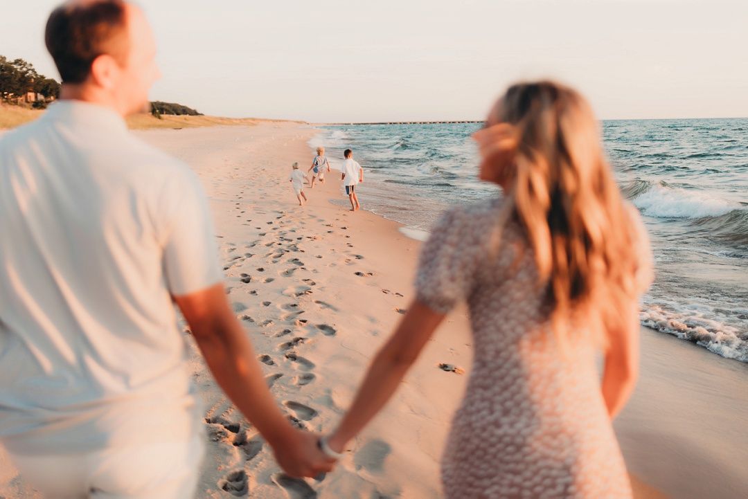 A couple holds hands walking on a beach at sunset while children play ahead of them.