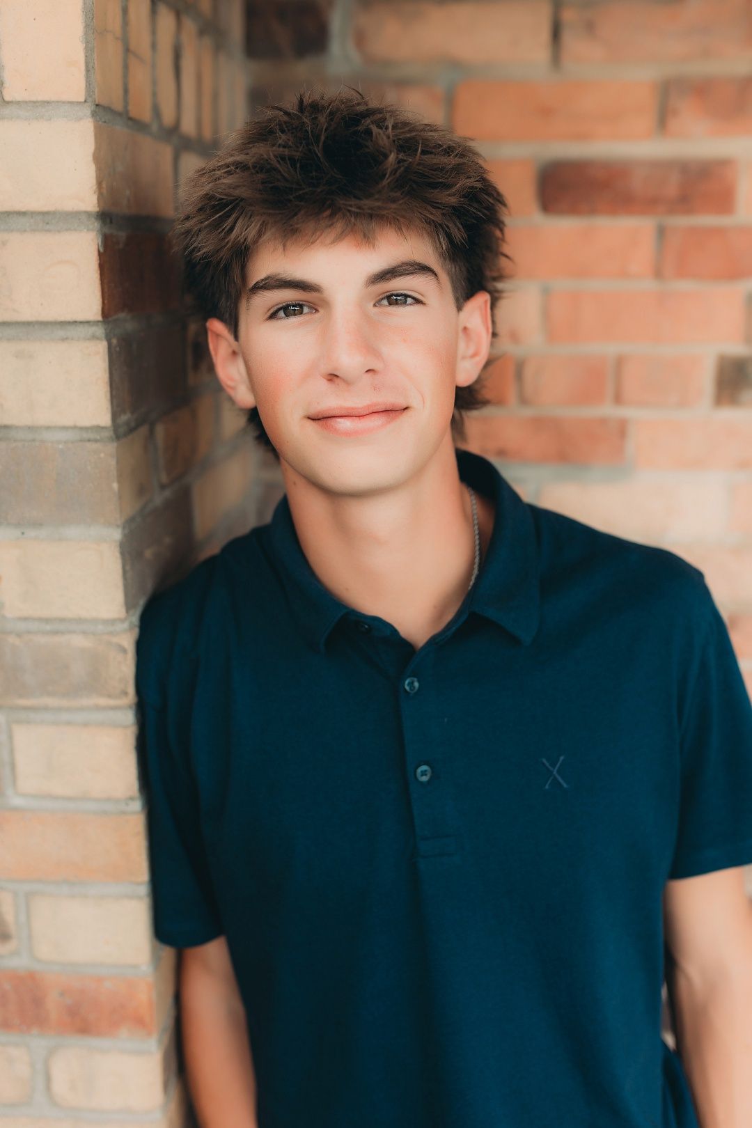 Teen boy in navy polo shirt leaning against brick wall smiling for senior portrait photo.