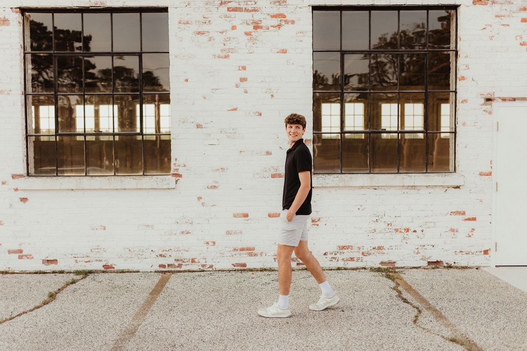 Young man in black shirt and shorts walking past white brick building.