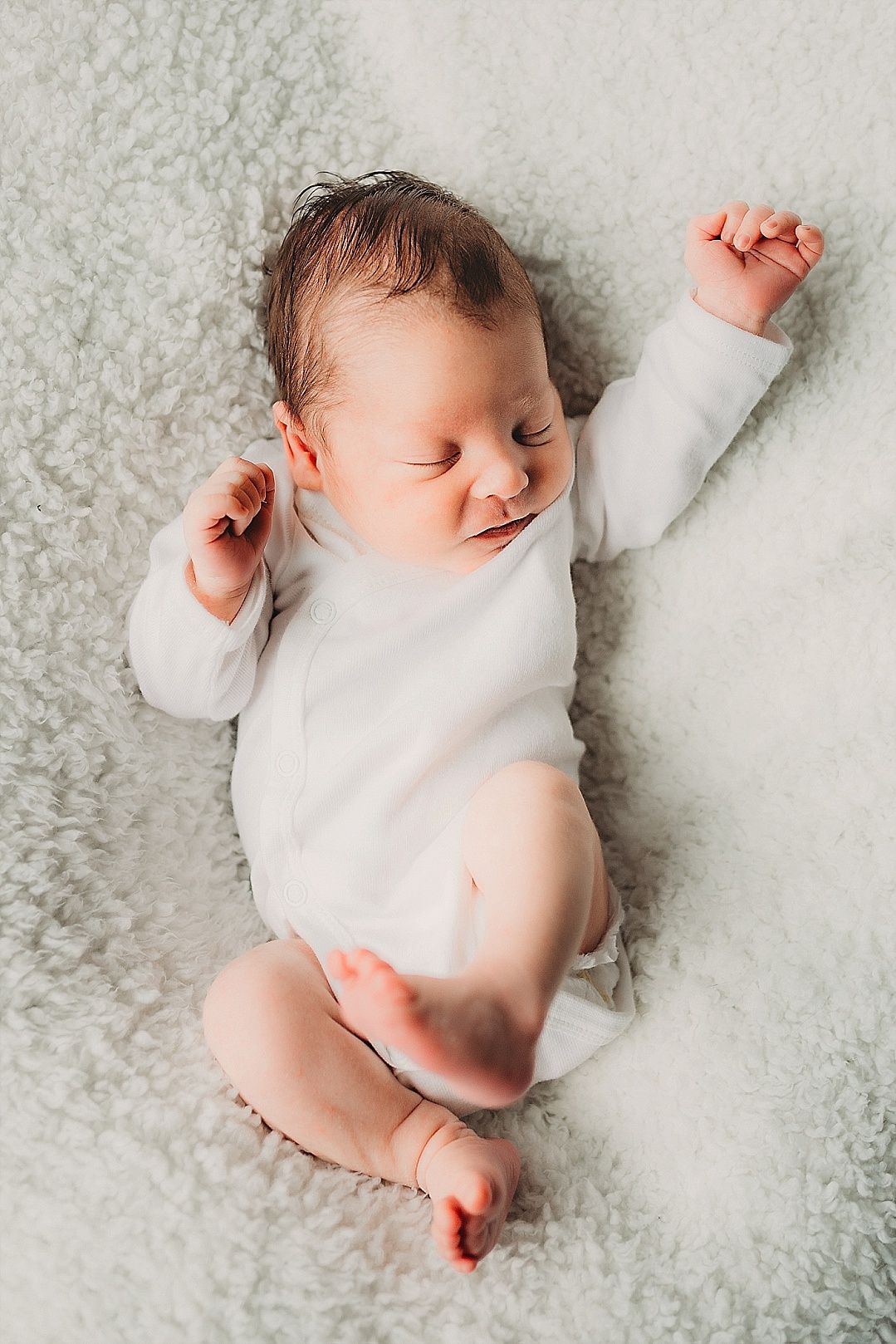 A peaceful newborn baby in a white onesie stretches arms up while lying on a fluffy blanket.