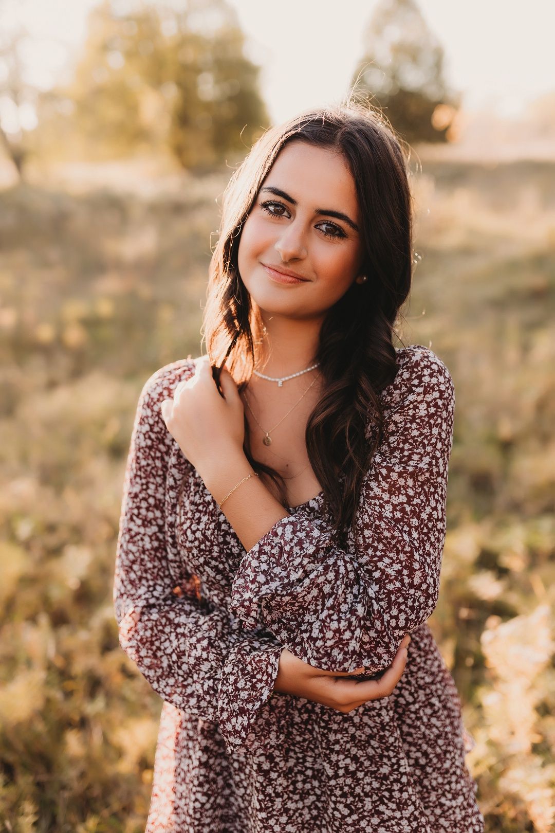 Brunette young woman in floral dress posing in golden sunlit field, hands crossed over chest.