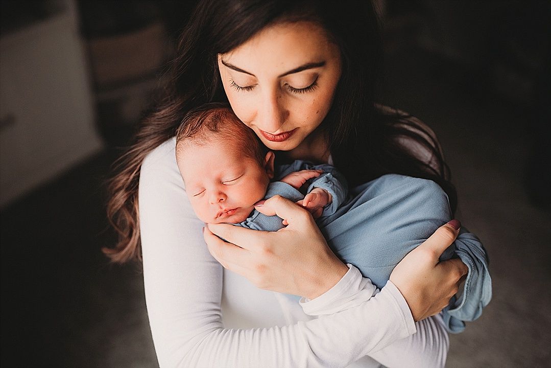 A mother tenderly cradles her sleeping newborn baby wrapped in a blue swaddle blanket.