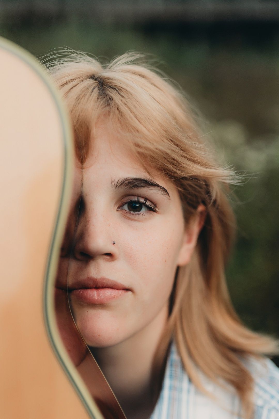 Close-up portrait of blonde teen girl with nose ring peeking over acoustic guitar body outdoors.