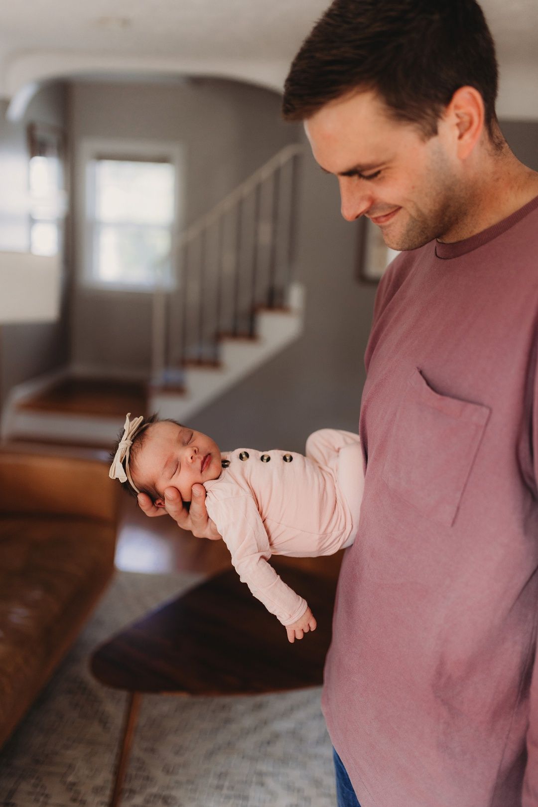 Smiling father in mauve shirt cradles sleeping newborn baby girl in pink outfit at home.