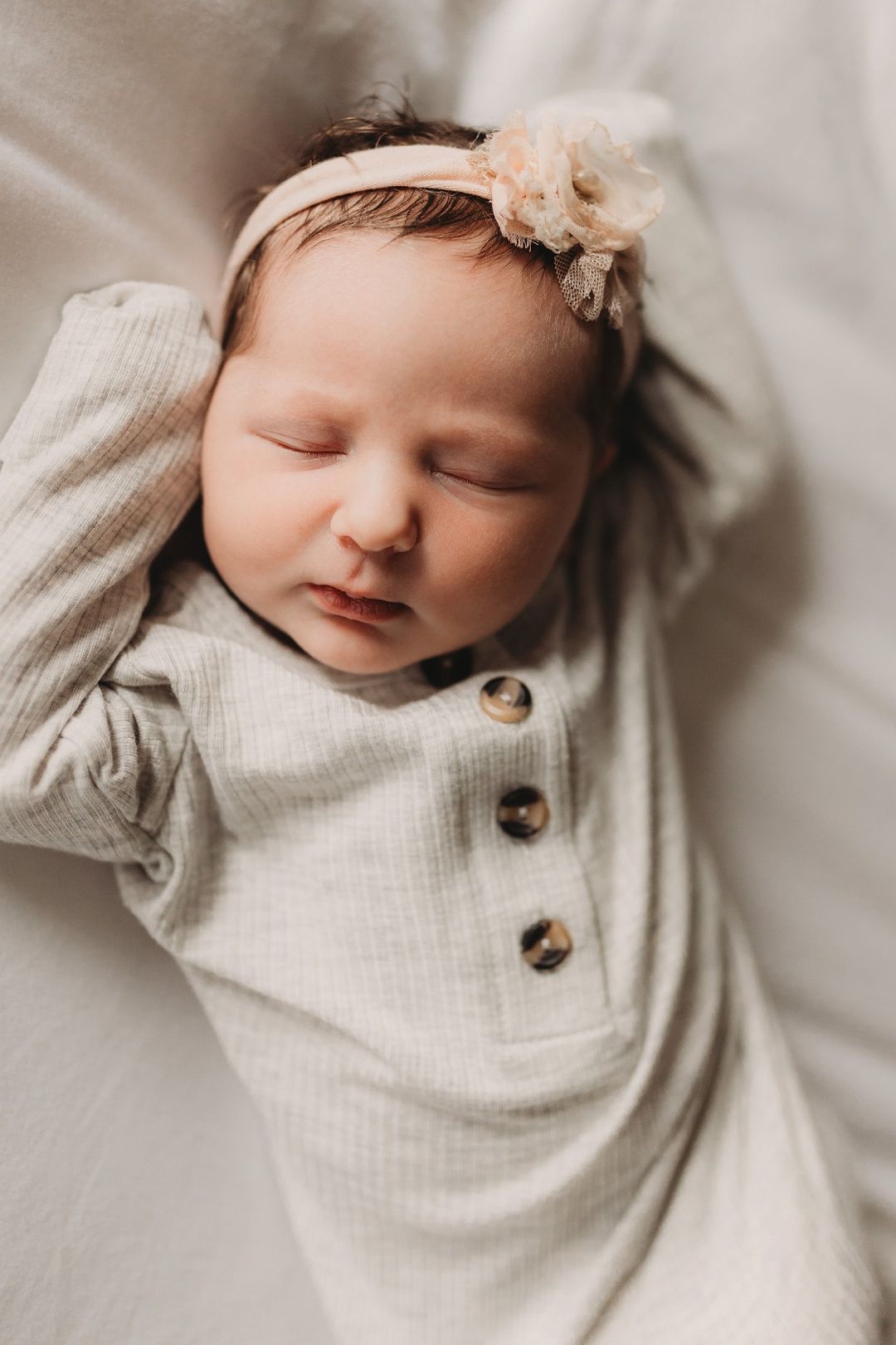Peaceful newborn baby girl in beige ribbed outfit and floral headband sleeping on soft blanket.