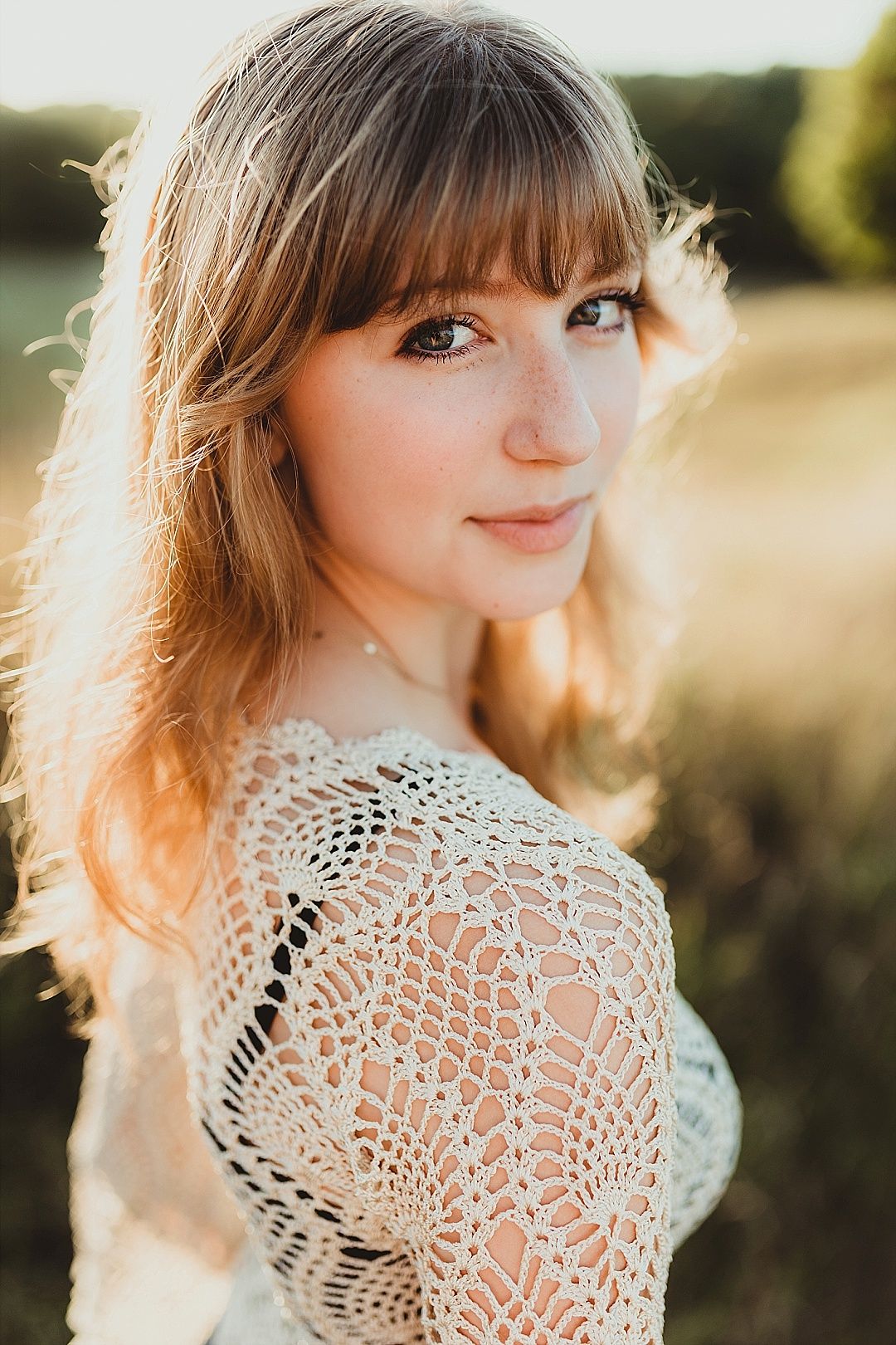 Young woman with bangs in crochet top smiling softly in golden sunlight outdoor field portrait.