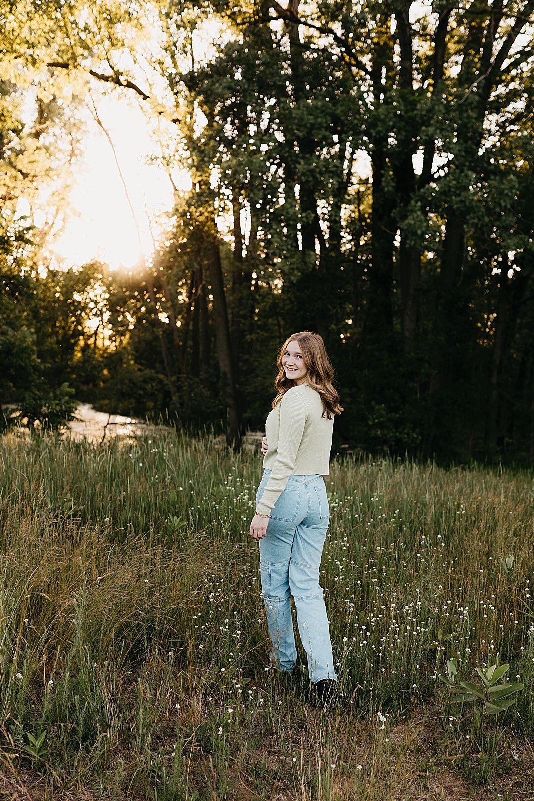 Smiling teenage girl in light sweater and jeans standing in a sunlit forest meadow.