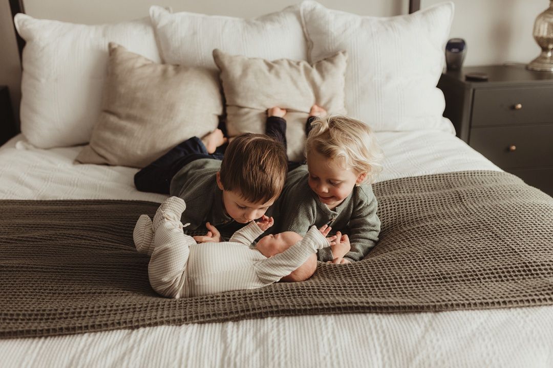 Two young siblings cuddle with their newborn baby on a cozy bed with neutral bedding.