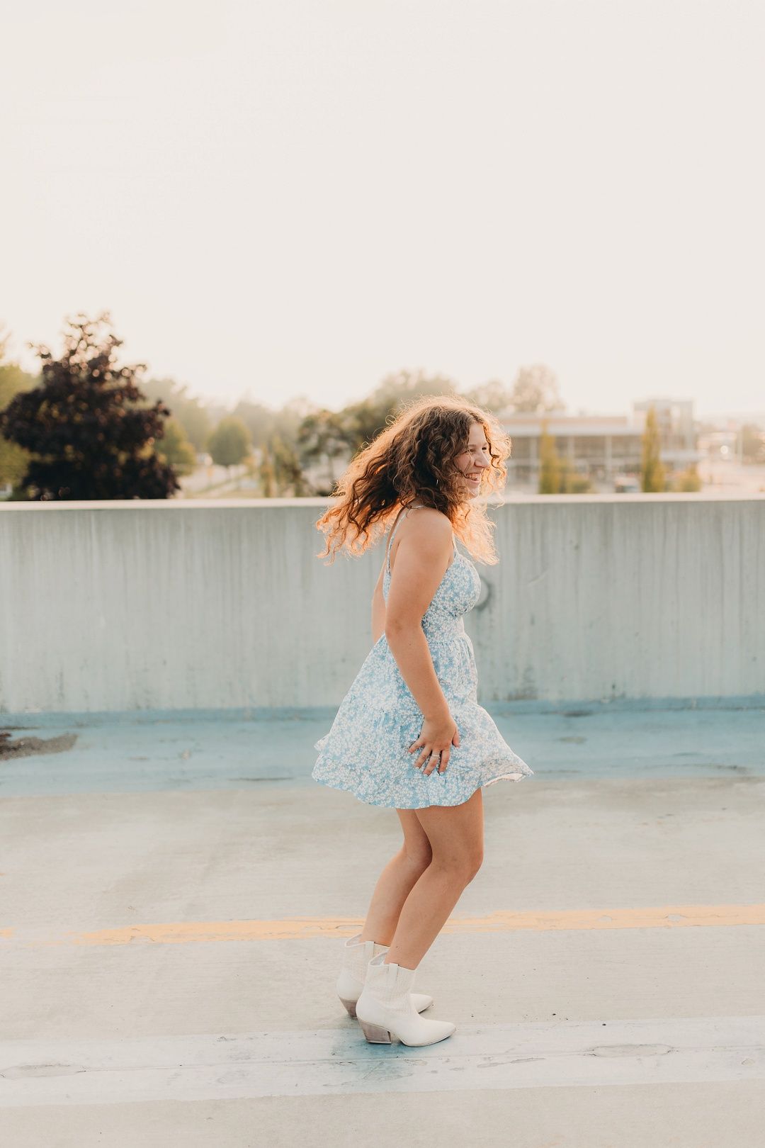 Young woman in floral sundress and white cowboy boots twirling on rooftop at golden hour.