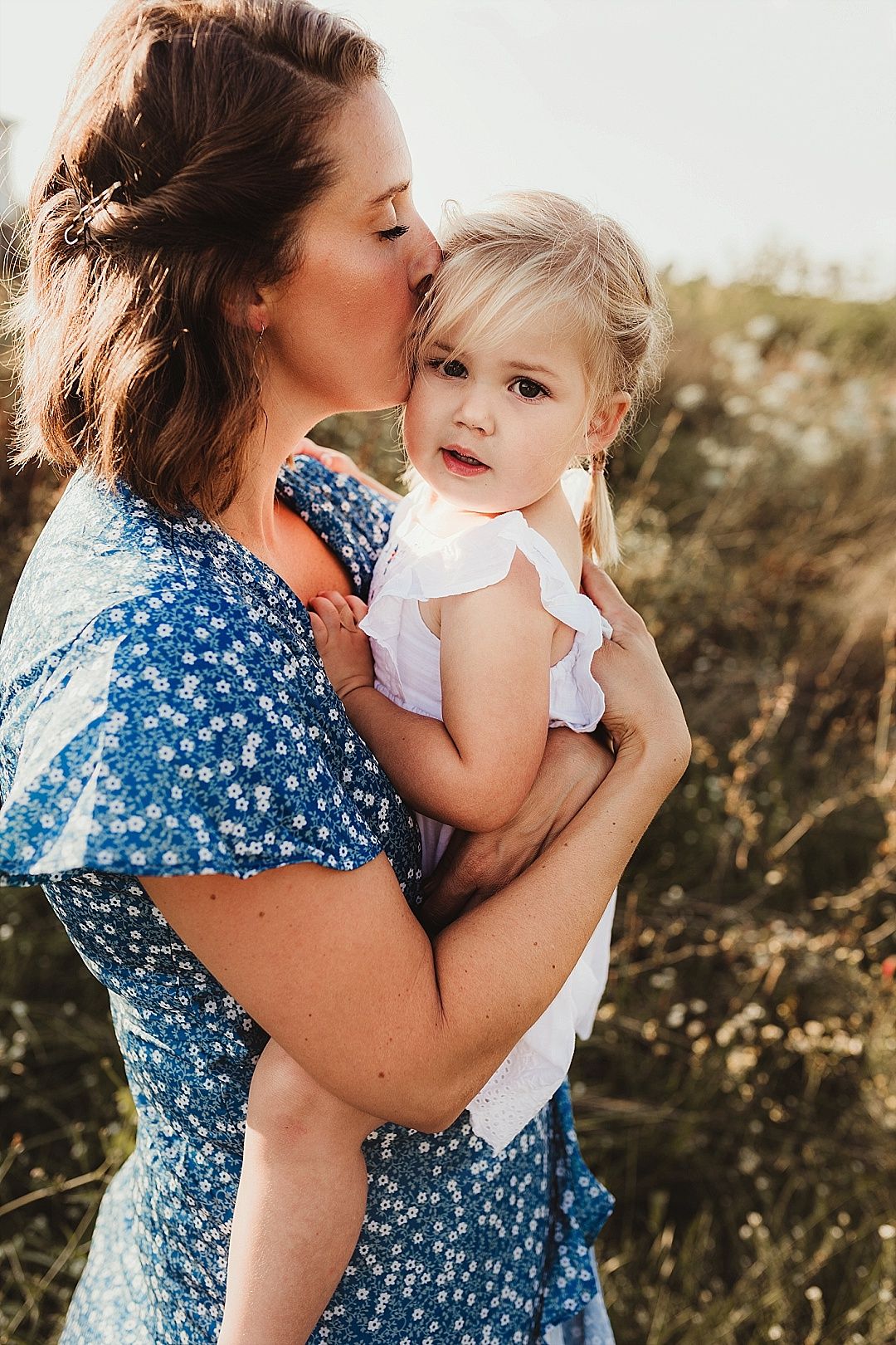 Mother in blue floral dress kissing blonde toddler daughter on the cheek outdoors.