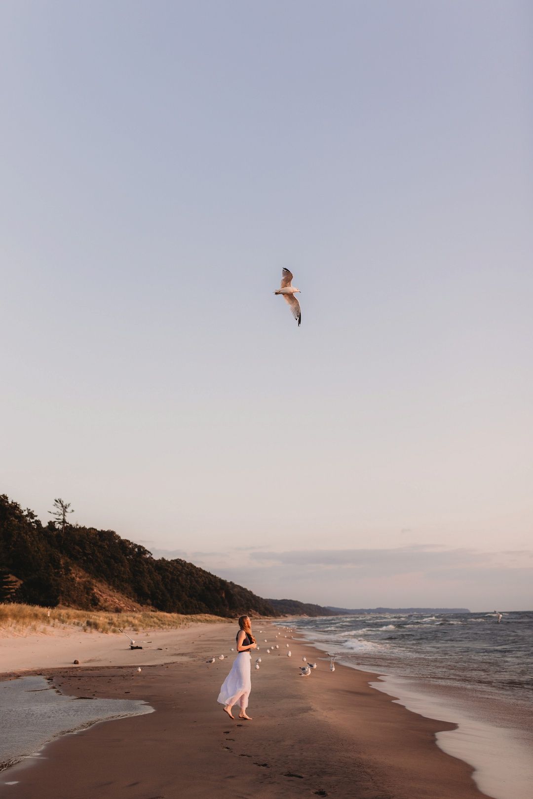 Woman in white dress walking on beach with seagull flying overhead.