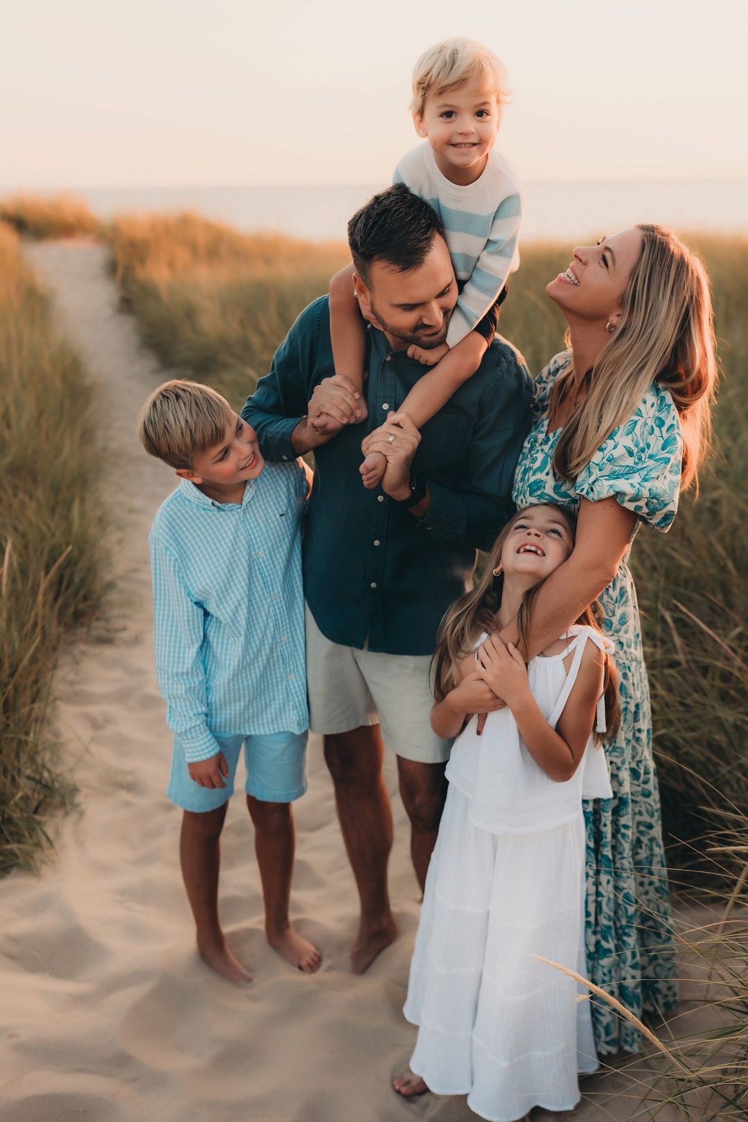 Joyful family of five posing on sandy beach path at golden sunset, parents with three children.