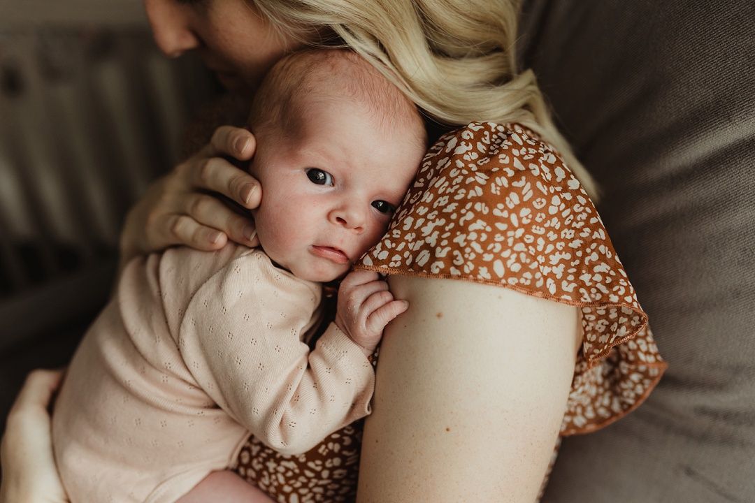 Blonde mother in floral dress tenderly holds sleeping newborn baby against her shoulder.