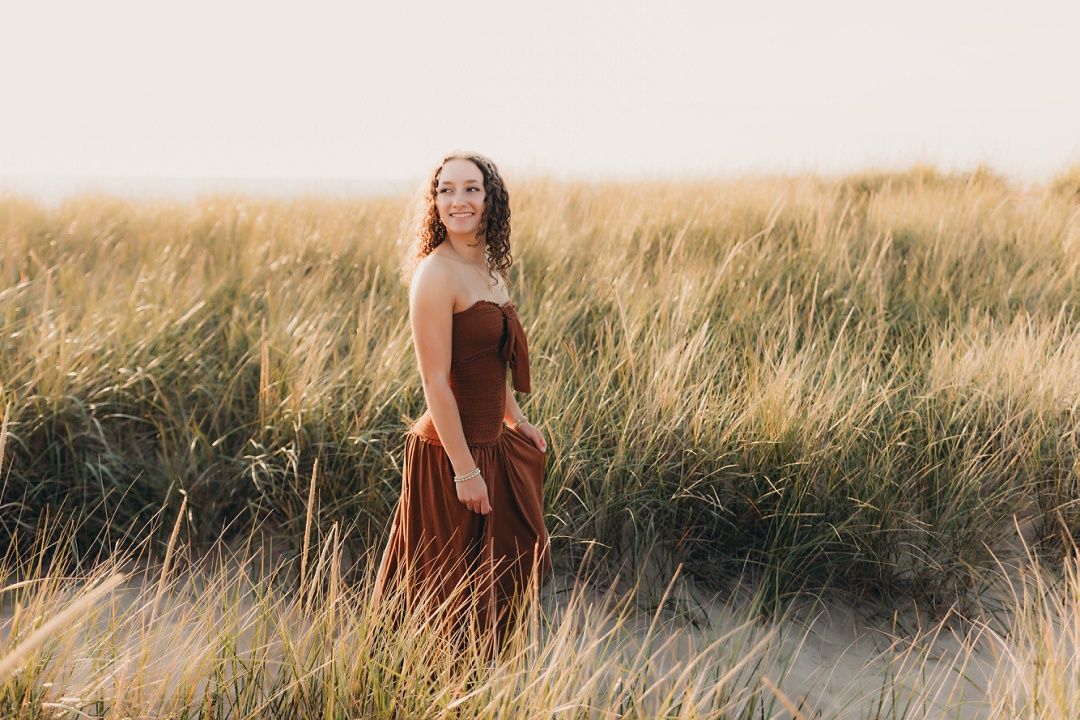 Young woman in rust strapless maxi dress standing among tall beach grass dunes at sunset.