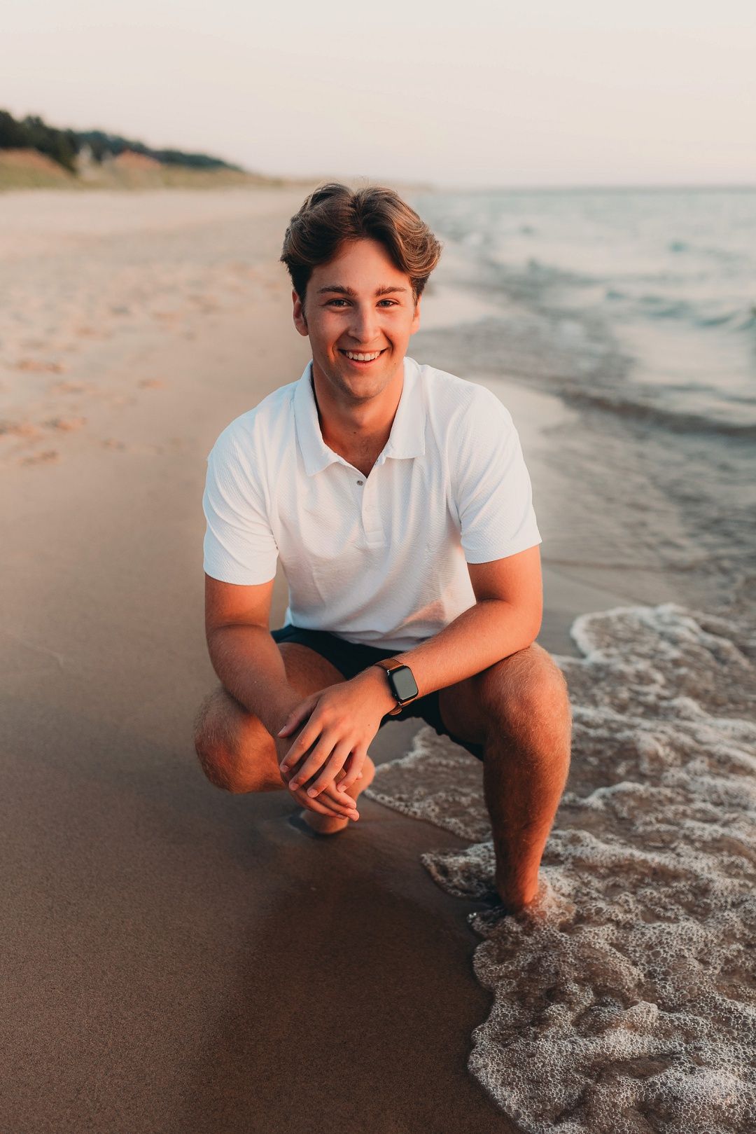 Smiling young man in white polo shirt crouching at the ocean shoreline during sunset.