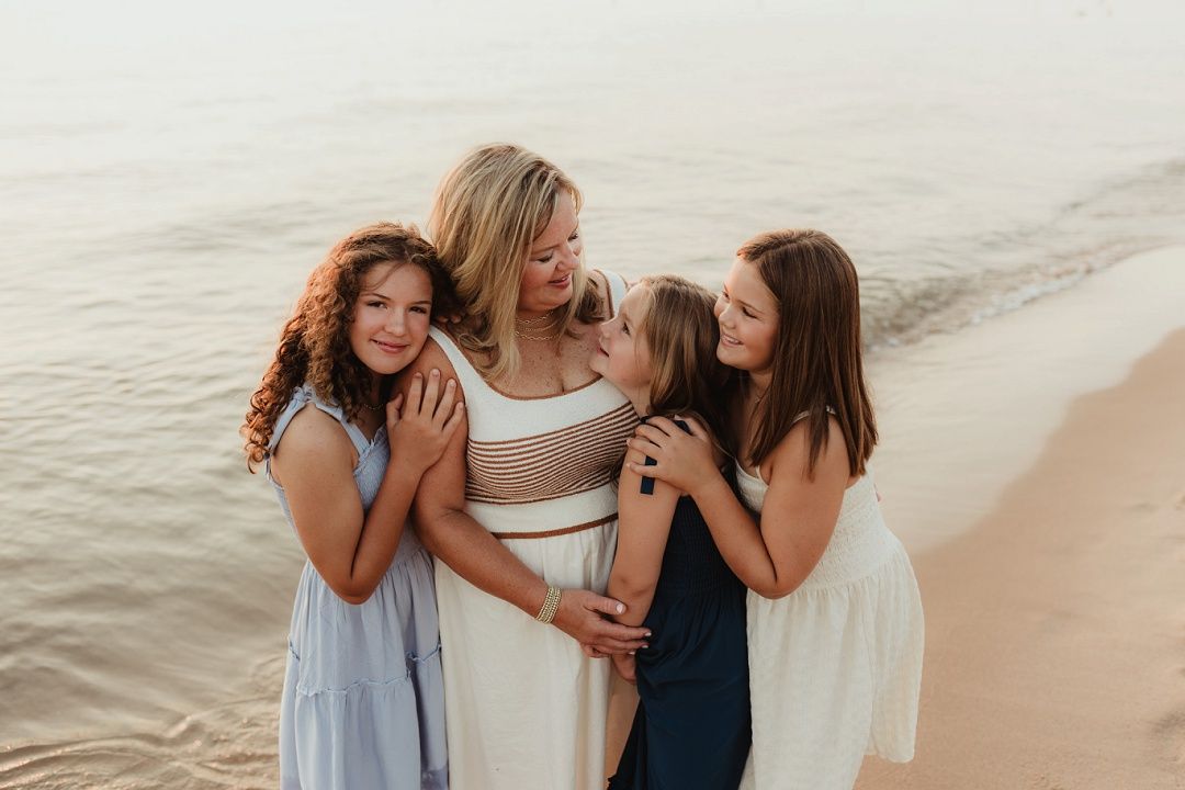 Mother embracing her three daughters on a serene sandy beach by the water.