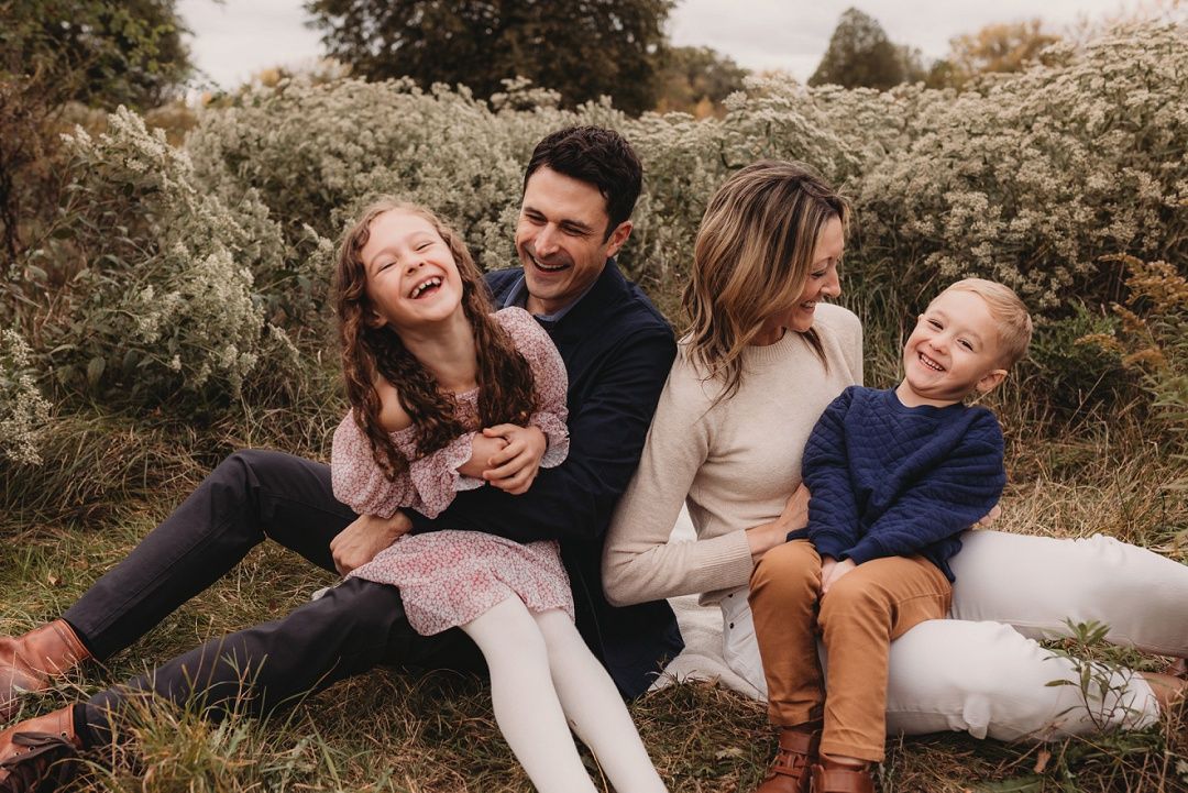 Happy family of four laughing together while sitting in a lush green outdoor setting.