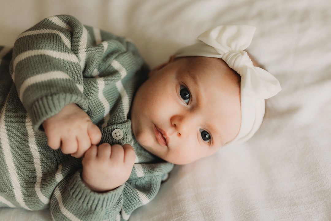 An alert baby with blue eyes wears a white bow headband and striped outfit.