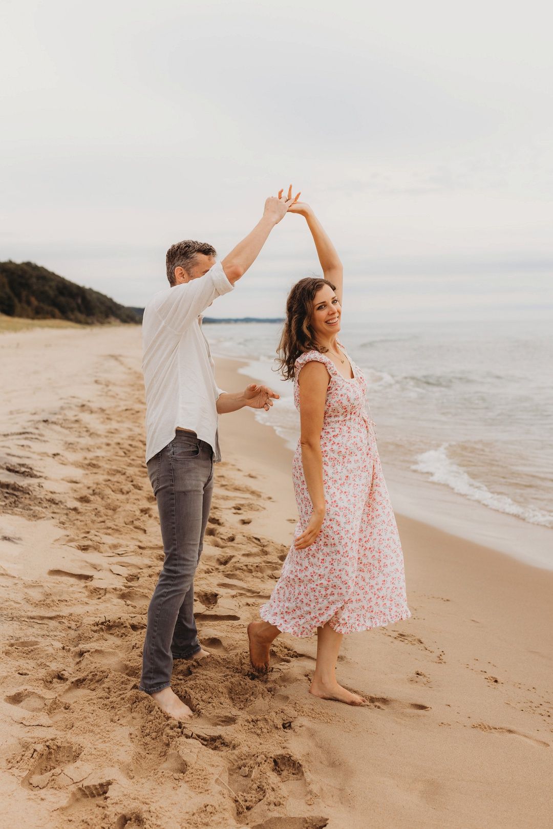 Couple dancing barefoot on sandy beach, man spinning woman in floral dress near ocean waves.