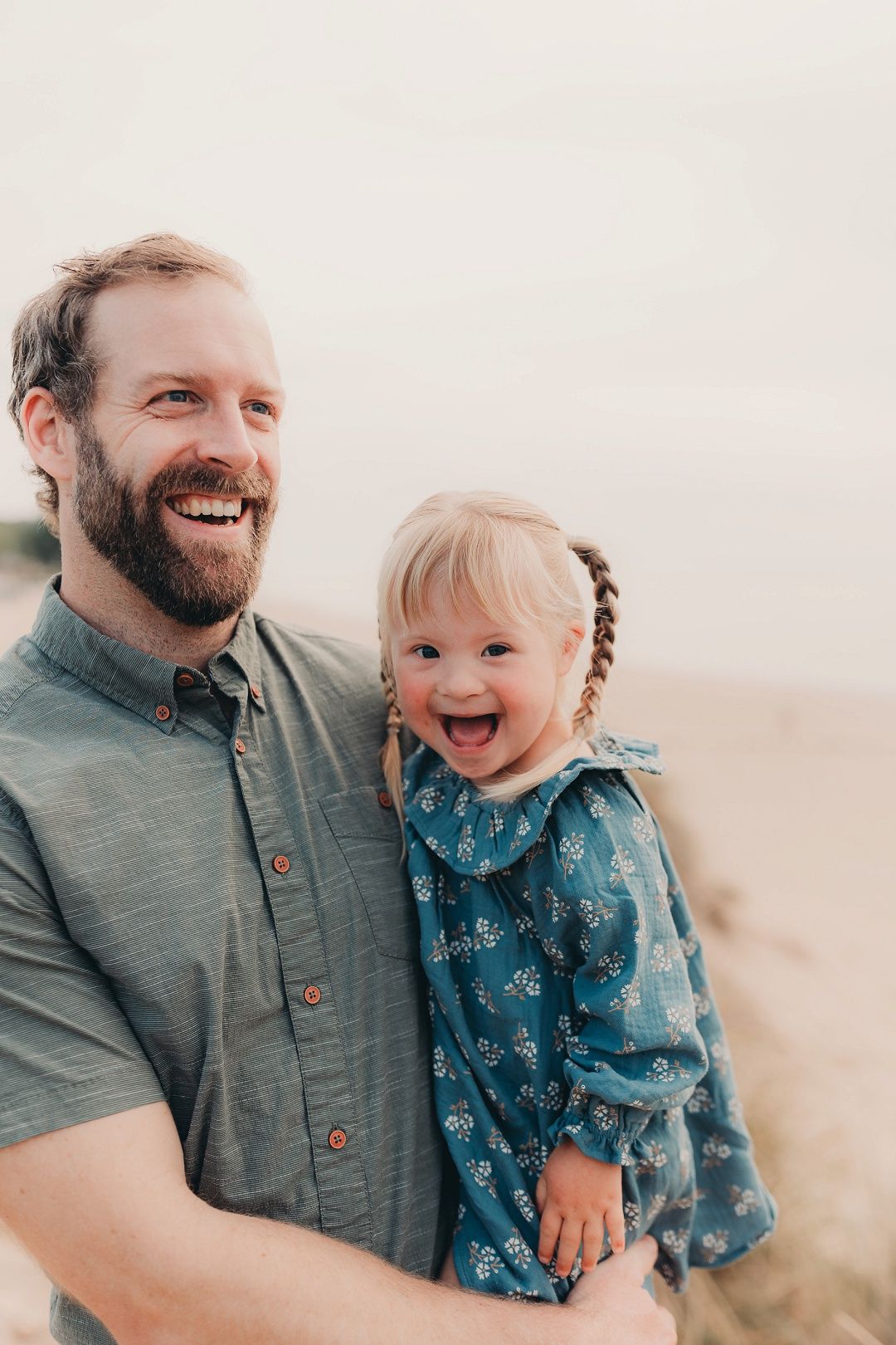 A bearded father laughs while holding his giggling toddler daughter outdoors near sandy dunes.