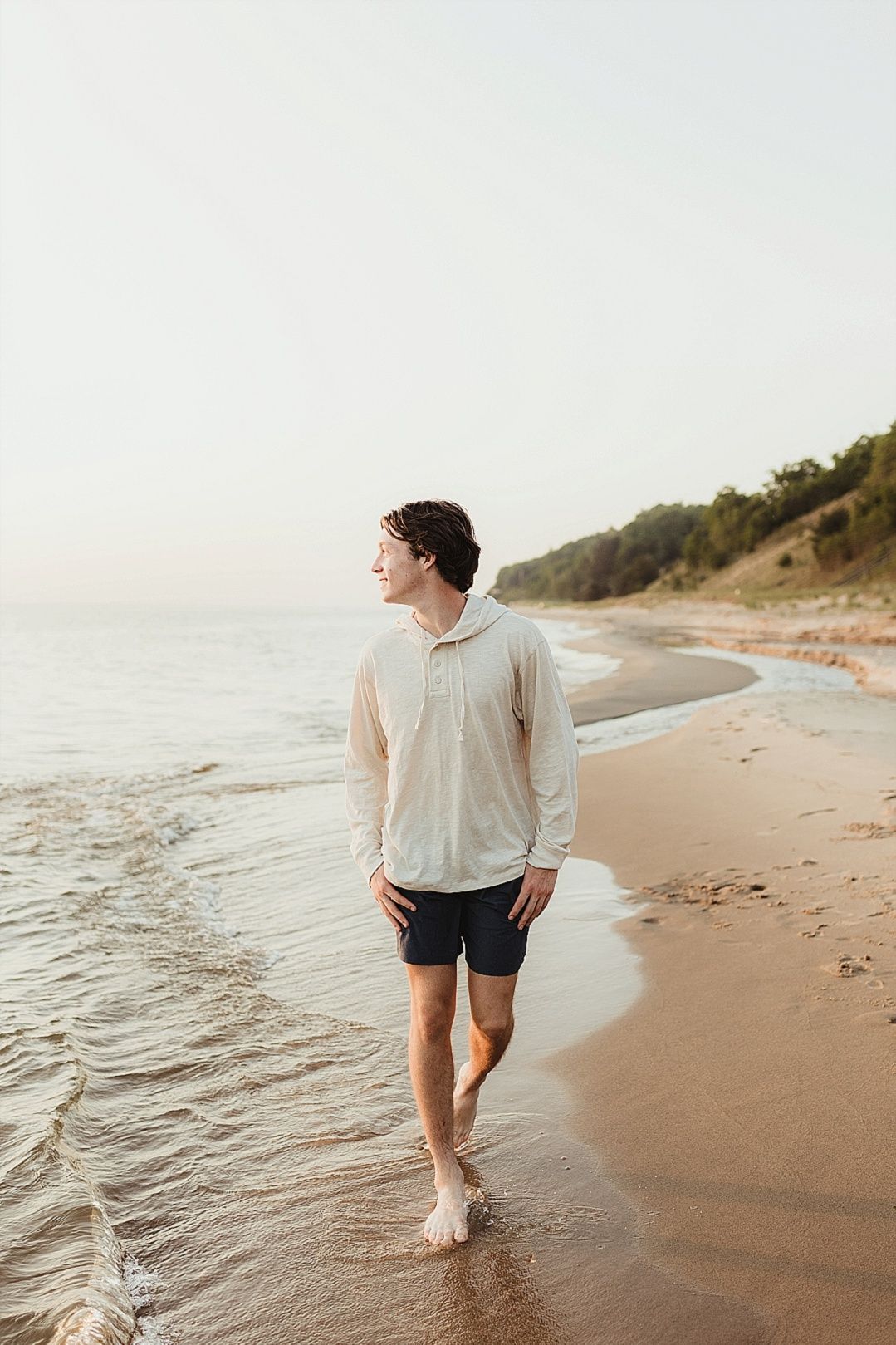 Young man in cream hoodie walking barefoot along a serene sandy beach at dusk.