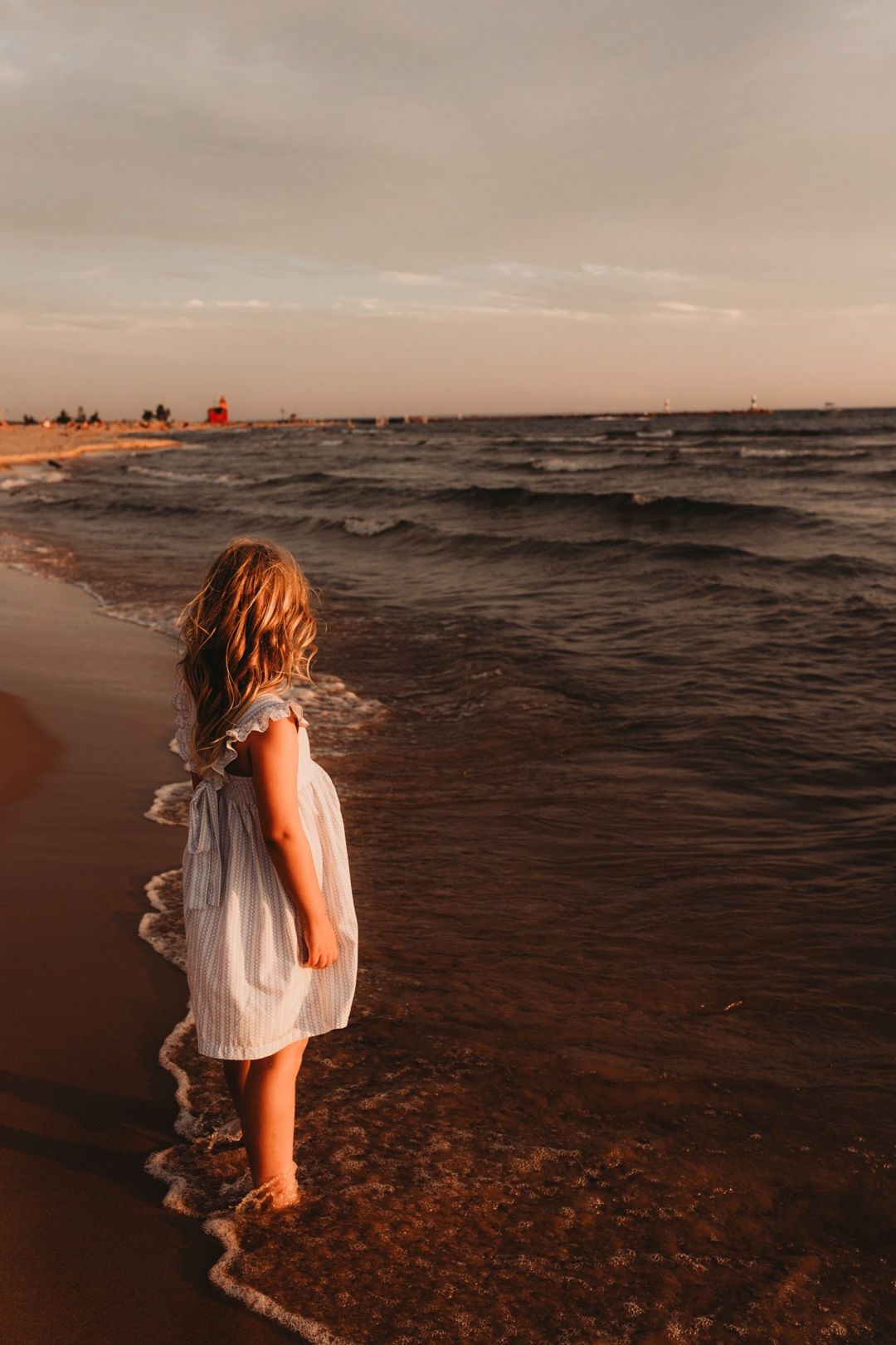 Young girl in white dress stands at ocean shoreline during golden sunset.