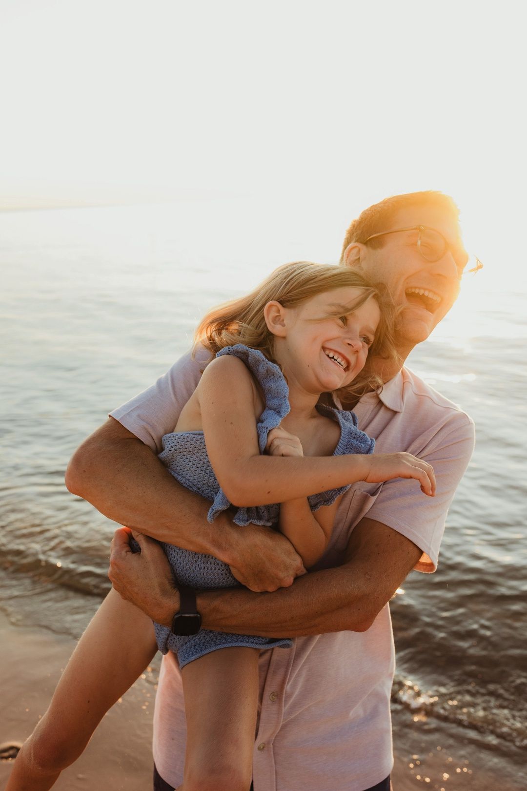 Father joyfully hugging laughing daughter on sunlit beach near ocean waves.