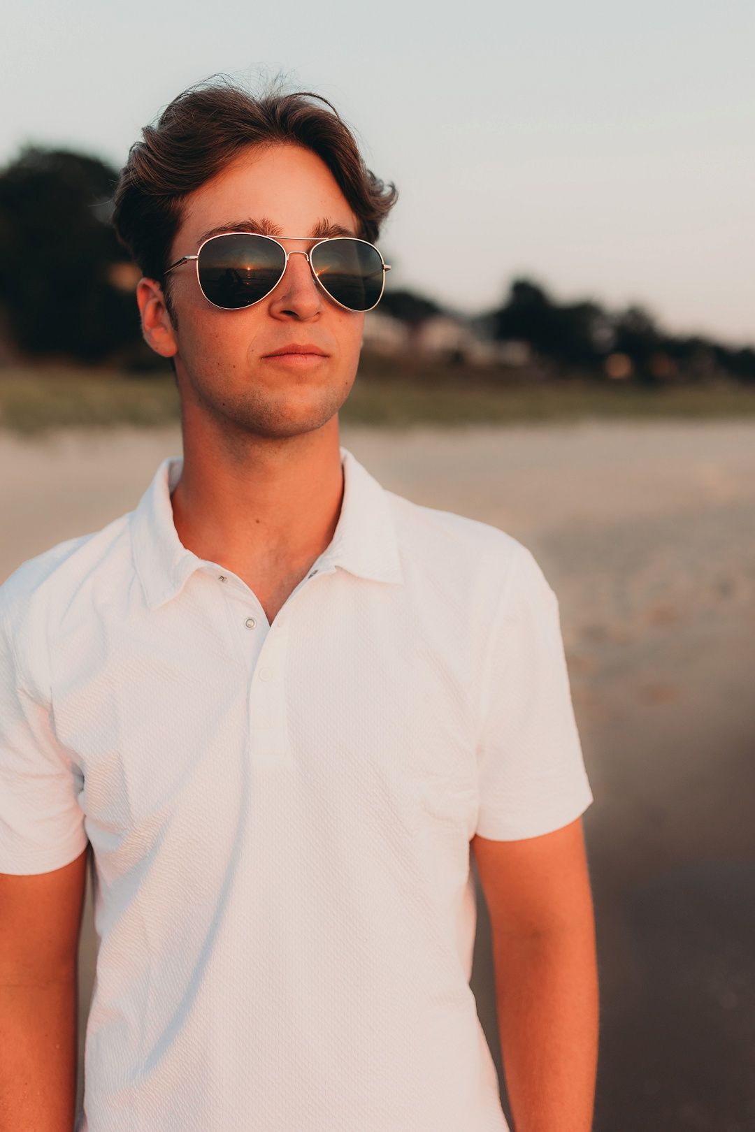Young man in white polo shirt wearing aviator sunglasses on beach.