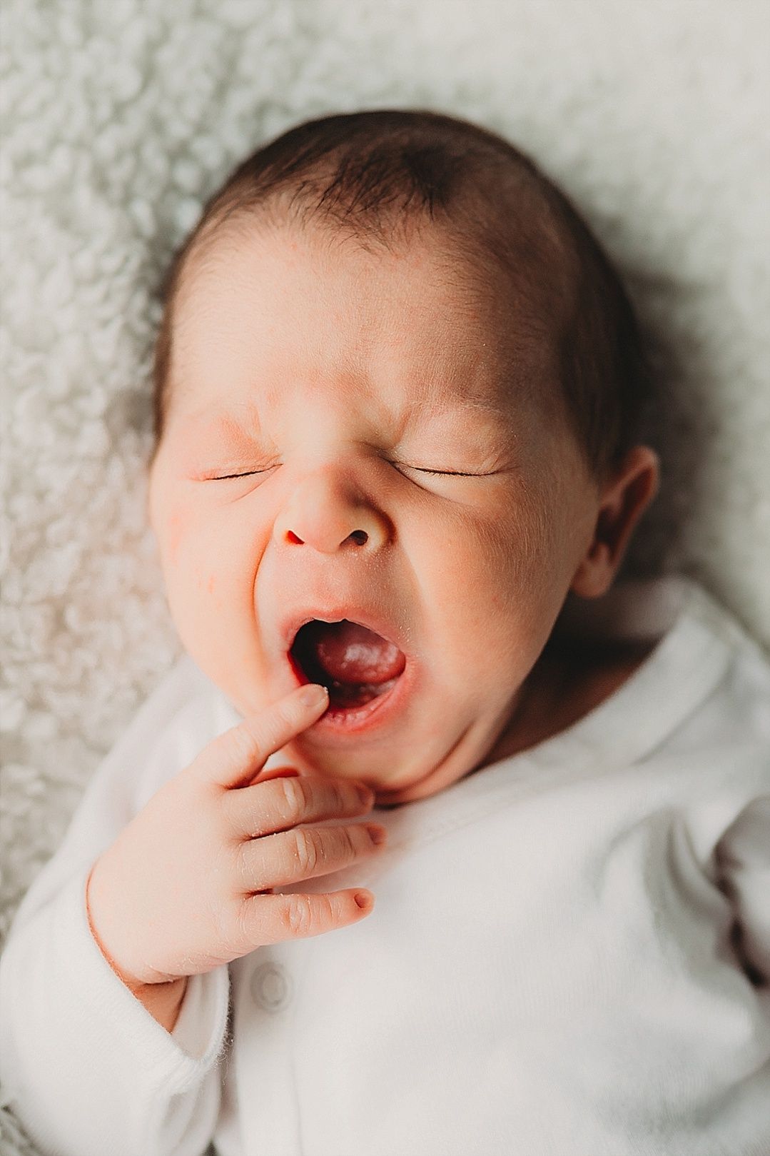 Close-up of a newborn baby yawning with hand near mouth while lying on a soft blanket.